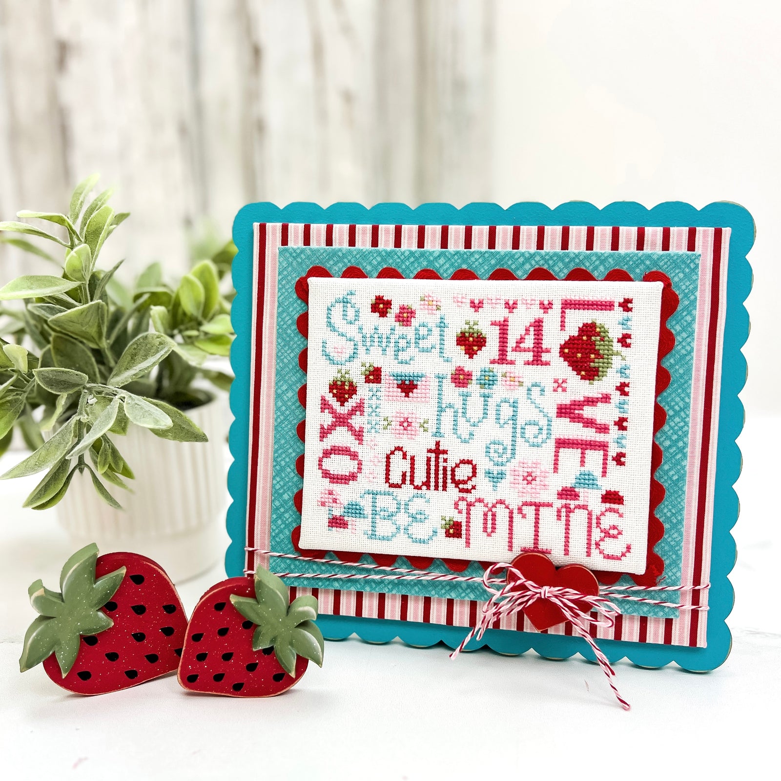 Painted scalloped wood cross stitch display with layered fabric backing, decorative trim, baker’s twine bow, and red heart button, designed for finishing and displaying completed cross stitch.