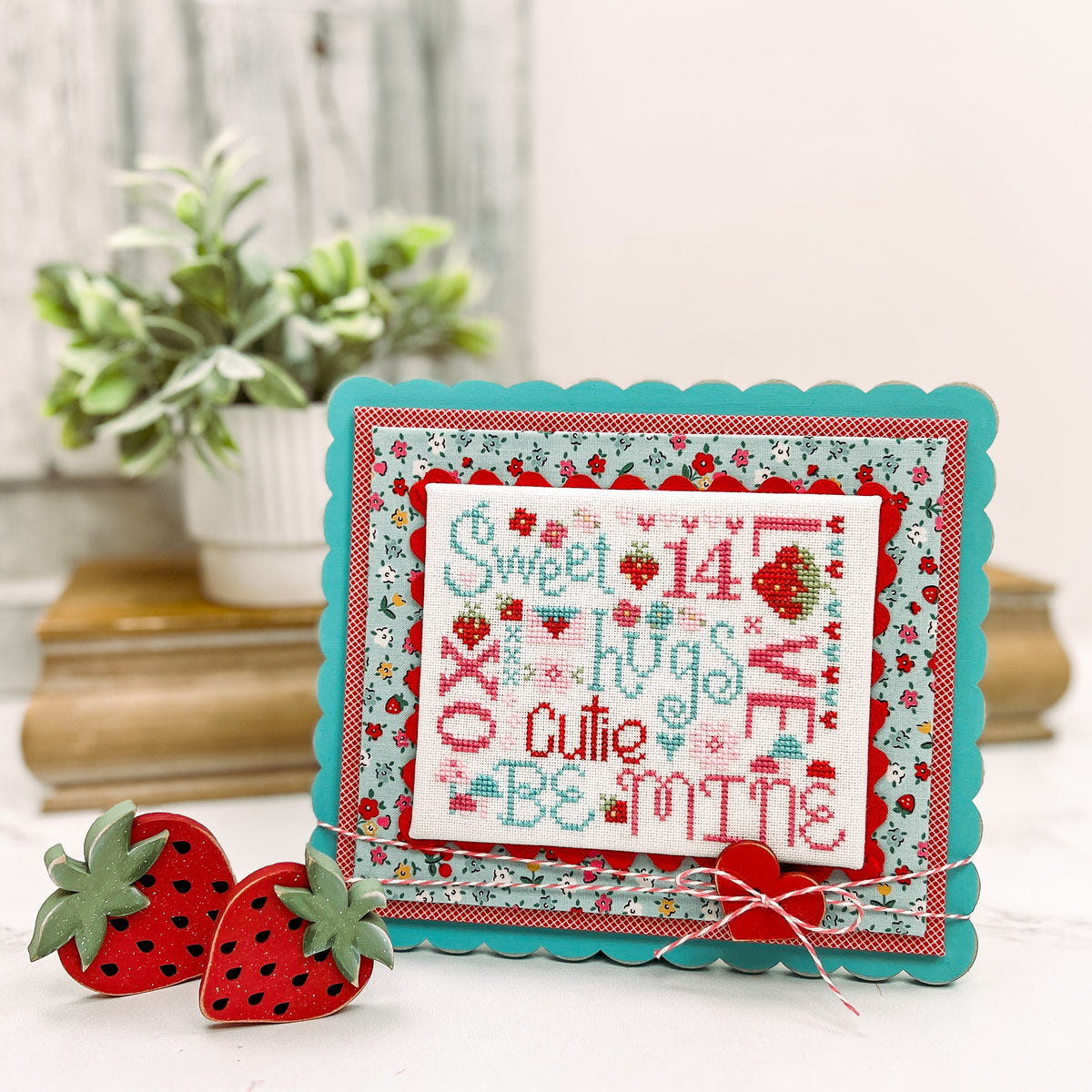 Painted scalloped wood cross stitch display with layered fabric backing, decorative trim, baker’s twine bow, and red heart button, designed for finishing and displaying completed cross stitch.