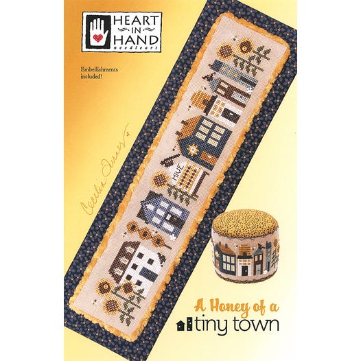 Cross-stitch pattern with houses, busy bees buzzing around a beehive and sunflowers on a yellow background, featuring &#39;Heart in Hand&#39; brand.