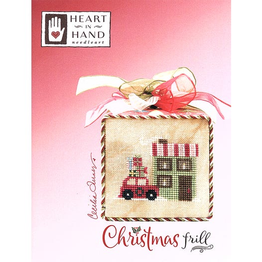 Cross-stitched design of a Christmas scene with a bow on a pink background, featuring 'Heart in Hand' brand.