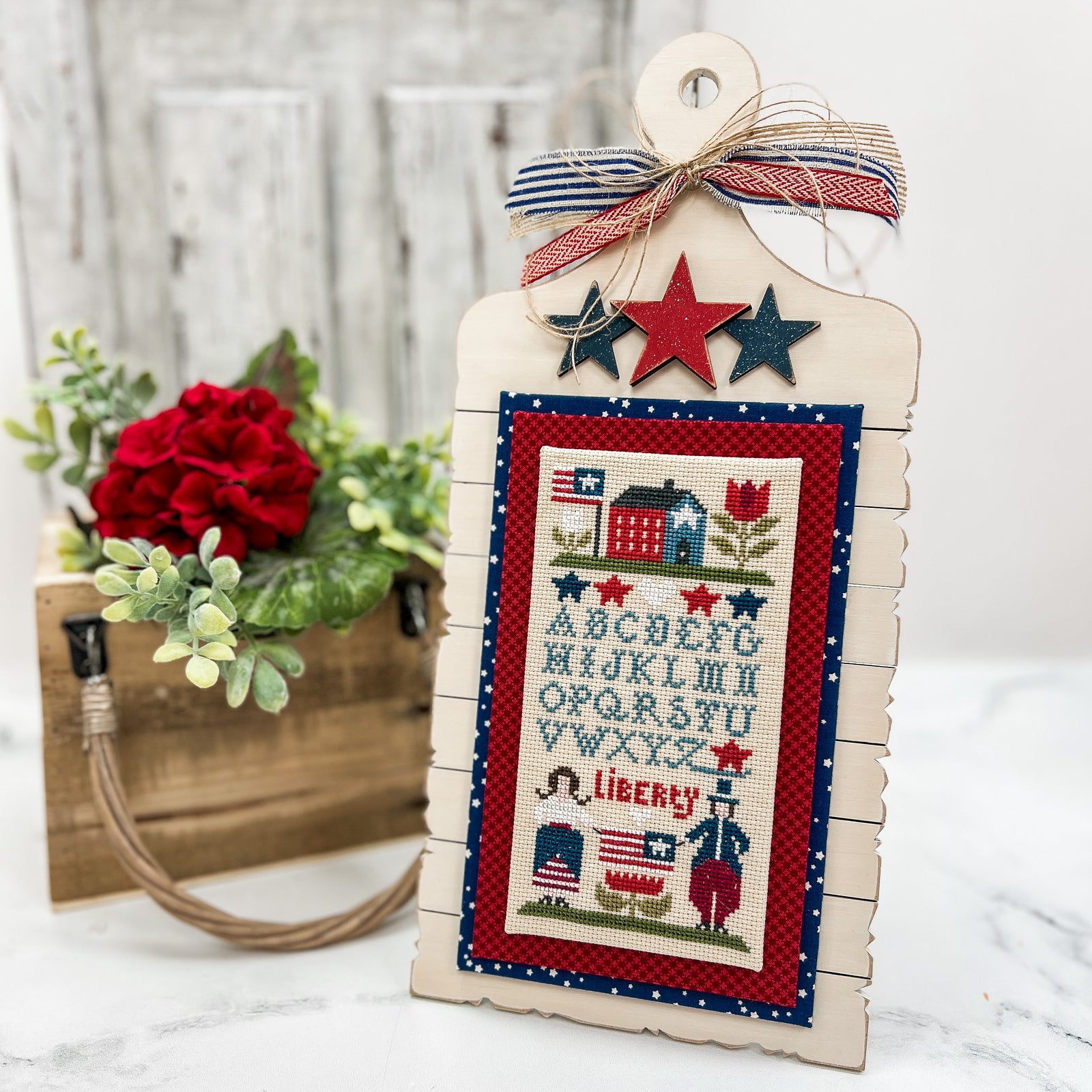 Pre-painted cream patriotic cross stitch display with red and blue wooden stars, holding an Americana cross stitch piece with red and blue fabric accents.