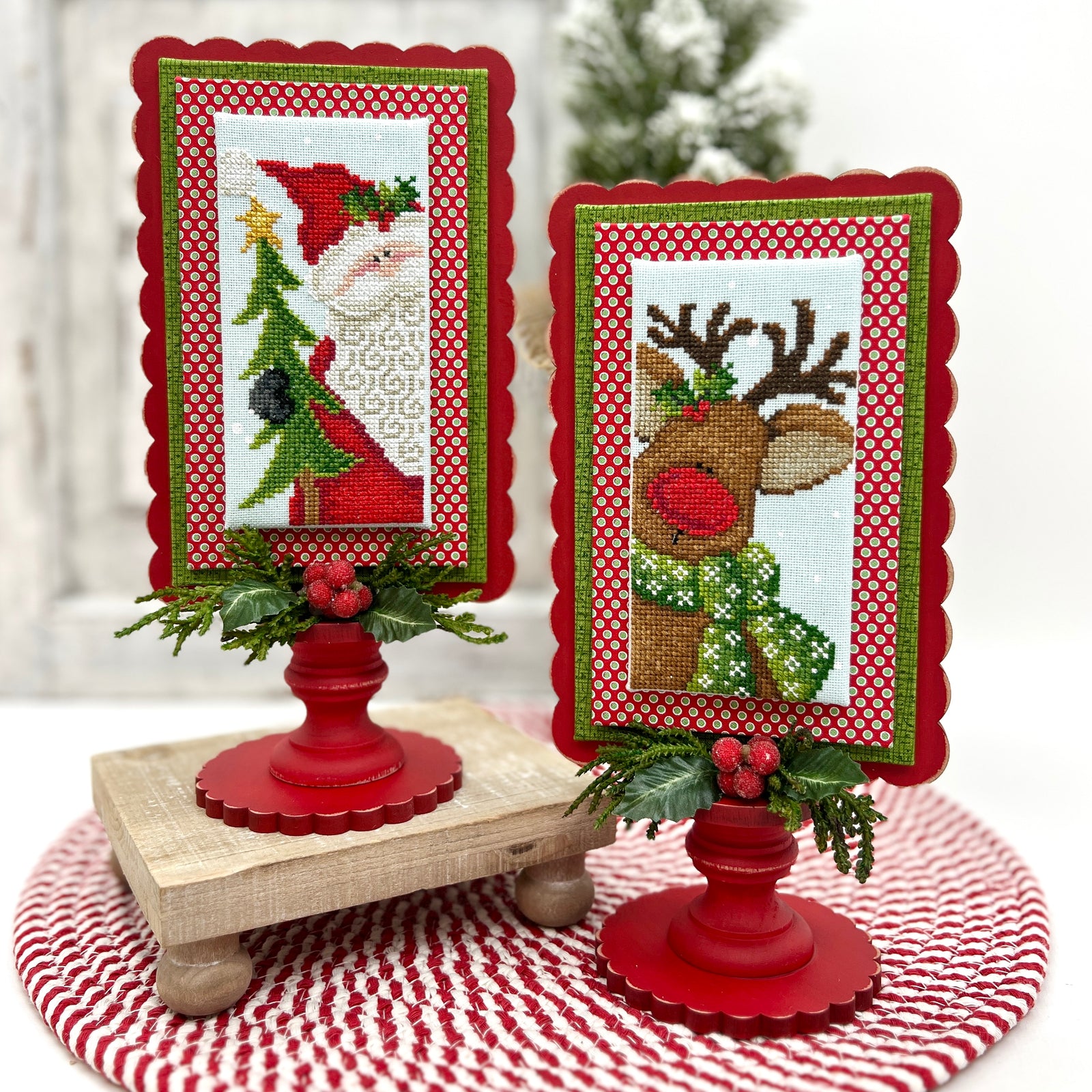 Two scalloped pedestal frames painted red, each displaying a festive cross stitch of Santa and a reindeer, decorated with greenery and berries beside a red “Merry” ornament.