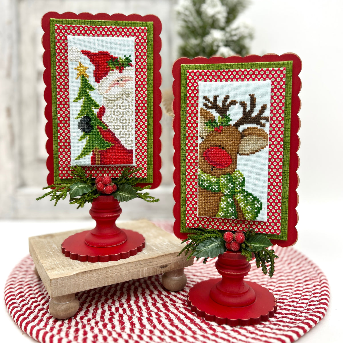 Two Christmas-themed cross-stitch frames on red stands with a blurred festive background.
