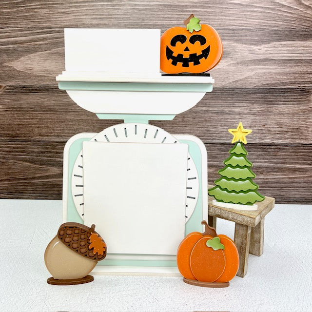 cross stitch display backer that looks like a vintage scale with fall and christmas seasonal shapes displayed with it