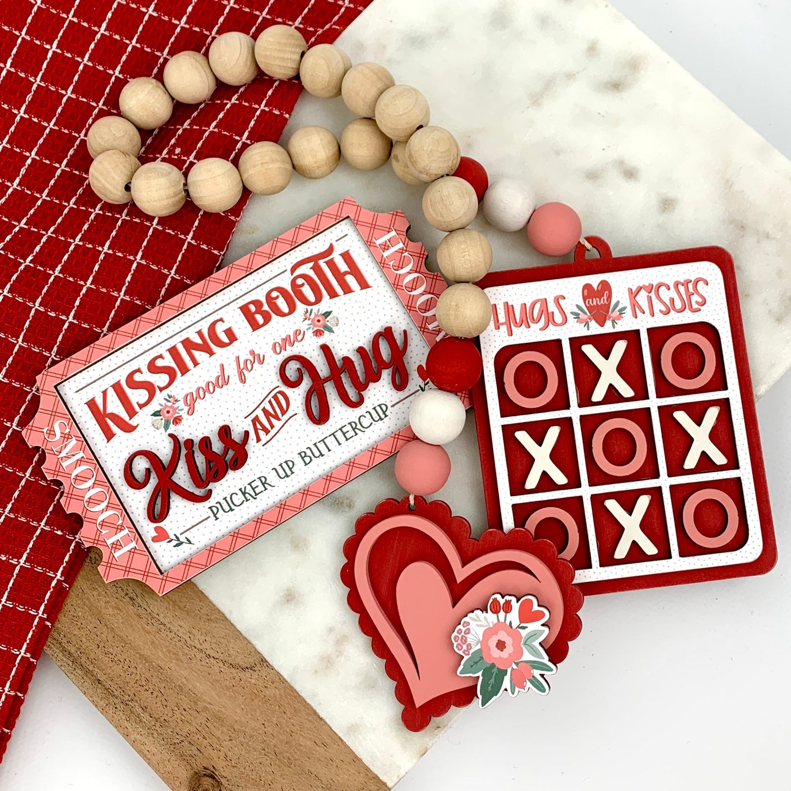 Red and pink Valentine wood decorations; kissing booth ticket and wood beaded garland with a layered heart and hugs and kisses tic tac toe.
